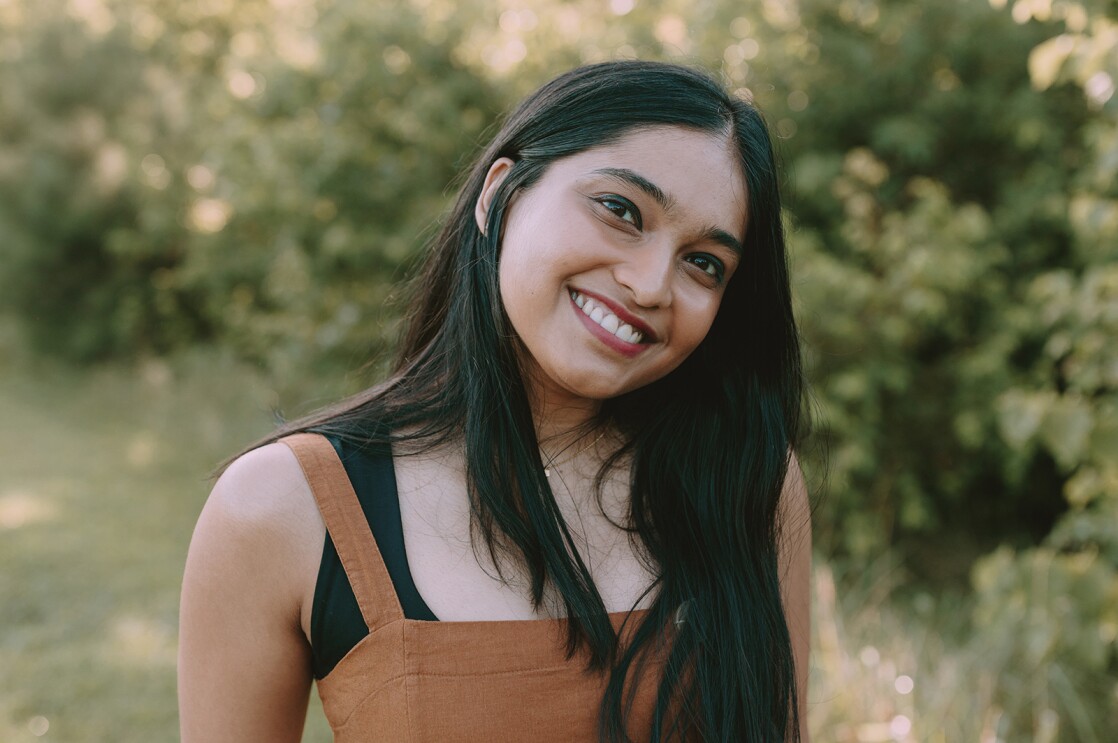 A headshot image of a woman standing in a green area with grass below her and a wall of bushes behind her.