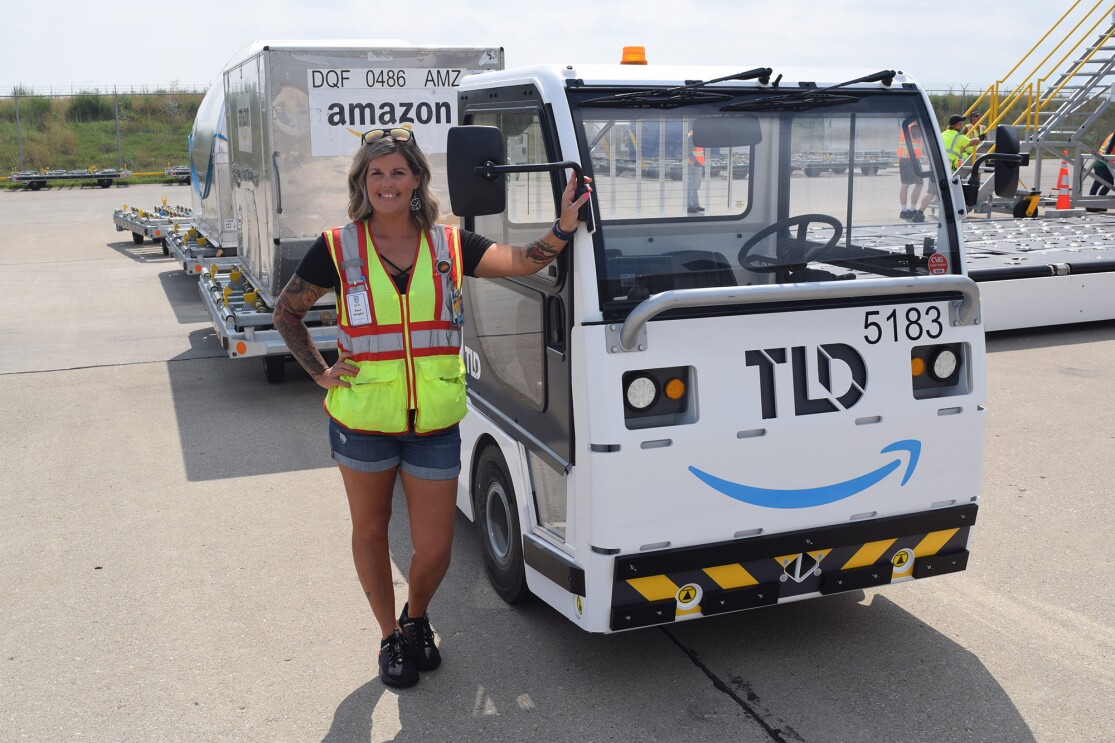 An image of a woman standing in front of a truck that is pulling large metal containers behind it. She is smiling for the photo and wearing a safety vest while working at the Amazon Air Hub in Kentucky.