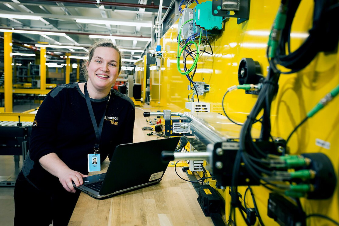Amazon Appretice, Nicola Elliott, leaning at her workbench with with her laptop. She is in the Bolton Fulfilment Centre next to a yellow wall with lots of wires and equipment attached to it. 