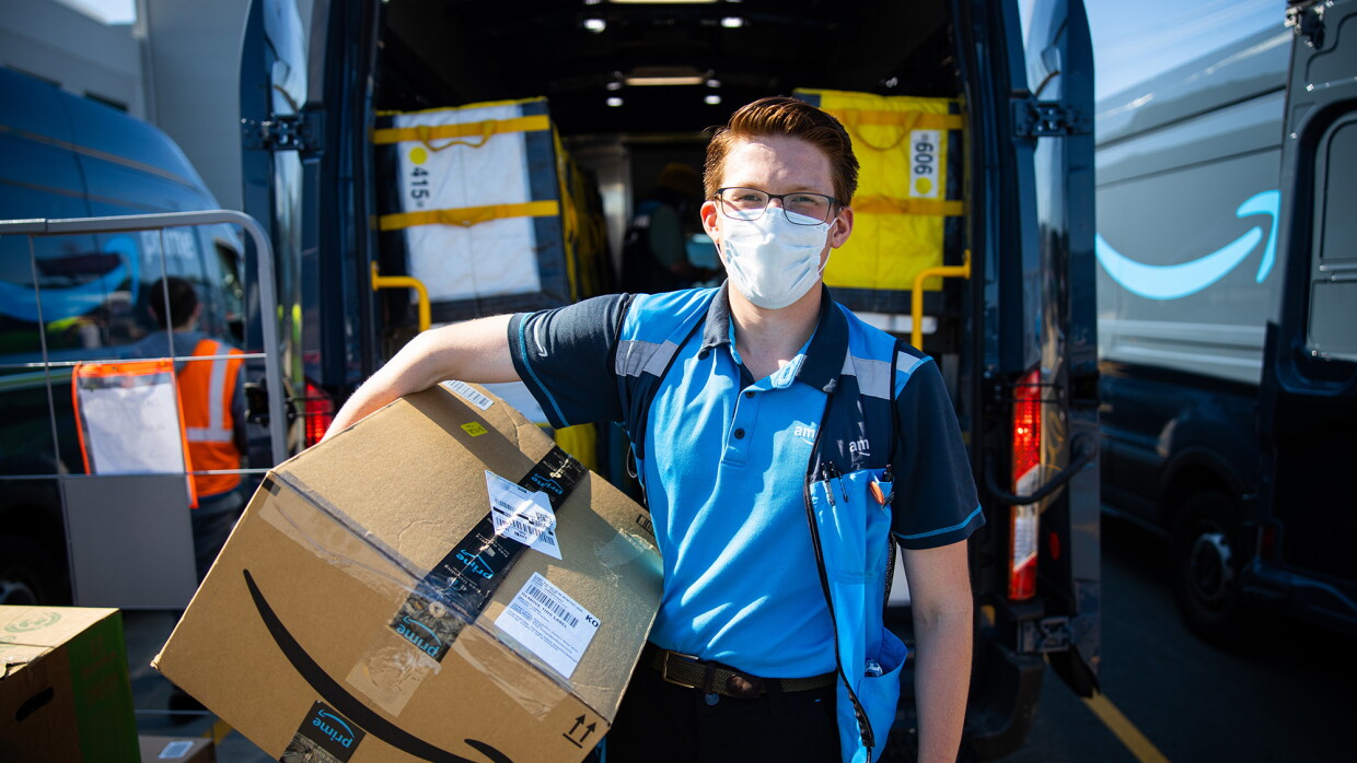 An Amazon delivery driver stands for a photo in front of his delivery van, holding a box and smiling under a mask. 