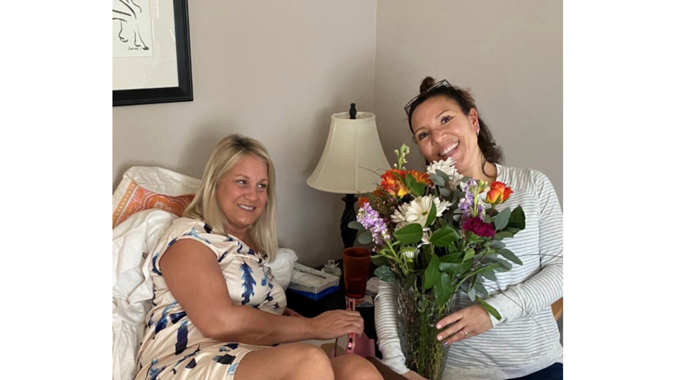 An image of two women smiling for a photo while sitting in a living room. One woman is holding flowers.