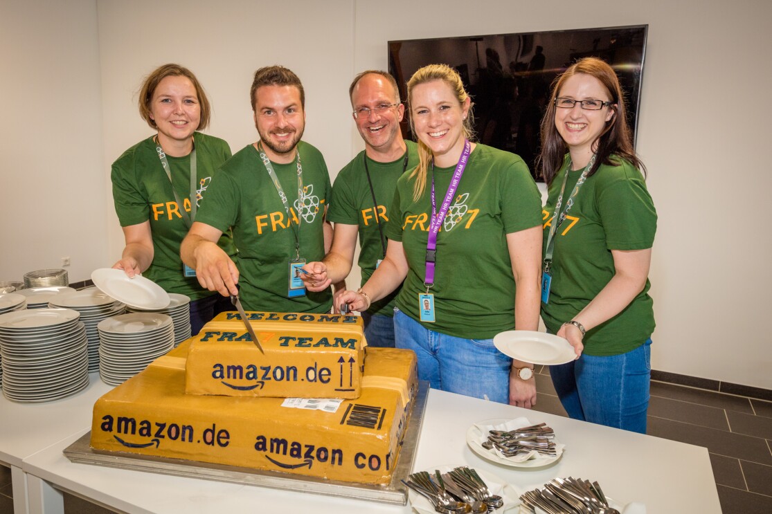 Ein zweistöckige Torte in Form von Päckchen trägt schwarze Beschriftungen mit "amazon.de" und "Fra7 Team". Die Torte steht auf einem Tisch auf dem ebenfalls viele Kuchenteller und Dessertgabeln angeordnet wurden. Fünf Mitarbeiter mit grünen T-Shirts sind um die Torte versammelt. Alle lächeln in die Kamera. Einer von ihnen: der Standortleiter. Er ist im Begriff, die Torte anzuschneiden.