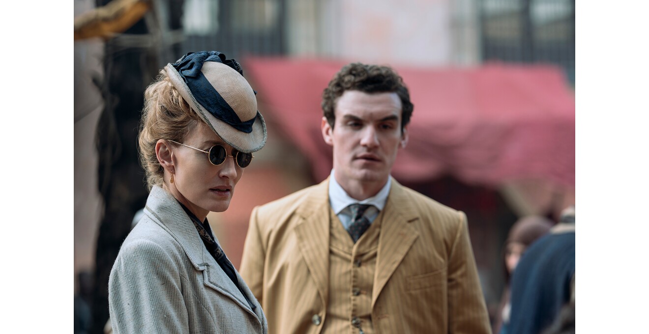 市場のような屋外で黒い丸眼鏡をかけた女性。リボンつきの帽子、ジャケットを着用。背後に三つ揃いスーツの男性が立っている。