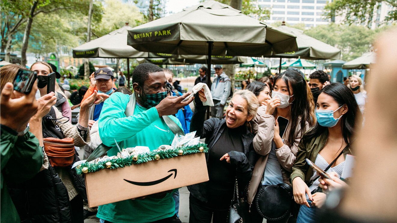 An image of a man passing out treats to an excited crowd. The box with the treats has the Amazon logo on it. The man is wearing a green Amazon shirt and a black and green checkered mask.