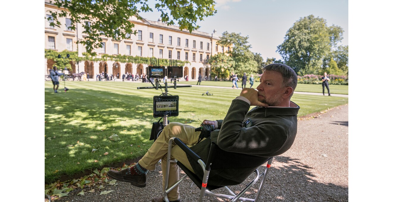 前庭に緑の芝生が広がる大きな建物。芝生の手前側の砂利道に置かれた監督椅子に座る男性。椅子の前には大小のモニターが3点設置されている。