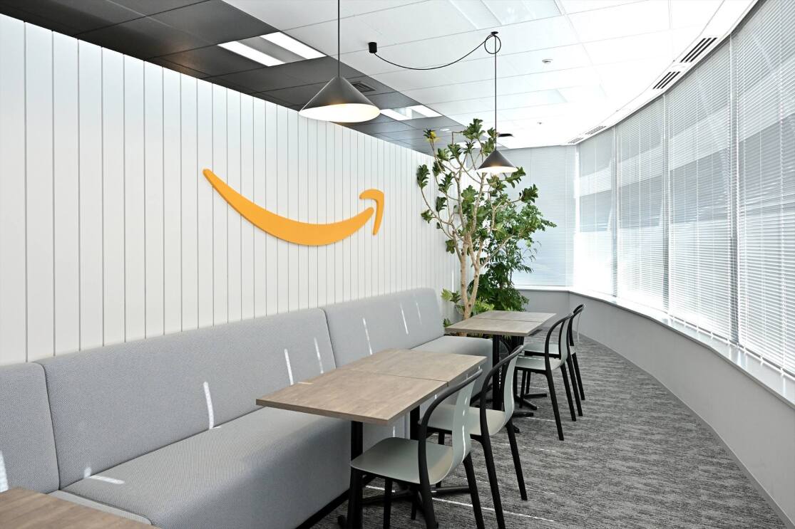 ミニマリストな空間にアマゾンロゴが白い壁に貼られている。壁の前に長いソファ、テーブル4台、そして椅子4脚が置かれている。