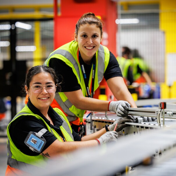 Dentro de un centro logístico, están dos mujeres arreglando una cadena metálica de transporte de paquetes. Las dos mujeres tiene el pelo oscuro largo y recogido en una cola. Las dos van en un camiseta negra de manga corta y un chaleco amarillo. Están las dos sonriendo , mirando a cámara, con las cuatro manos y agarrando algo y una de ellas agarra un destornillador.