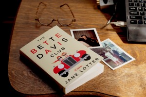 A copy of The Bette Davis Club sits on a tabletop, on top of vintage photographs. In the background rests a pair of glasses, to the right of the book is a laptop computer.