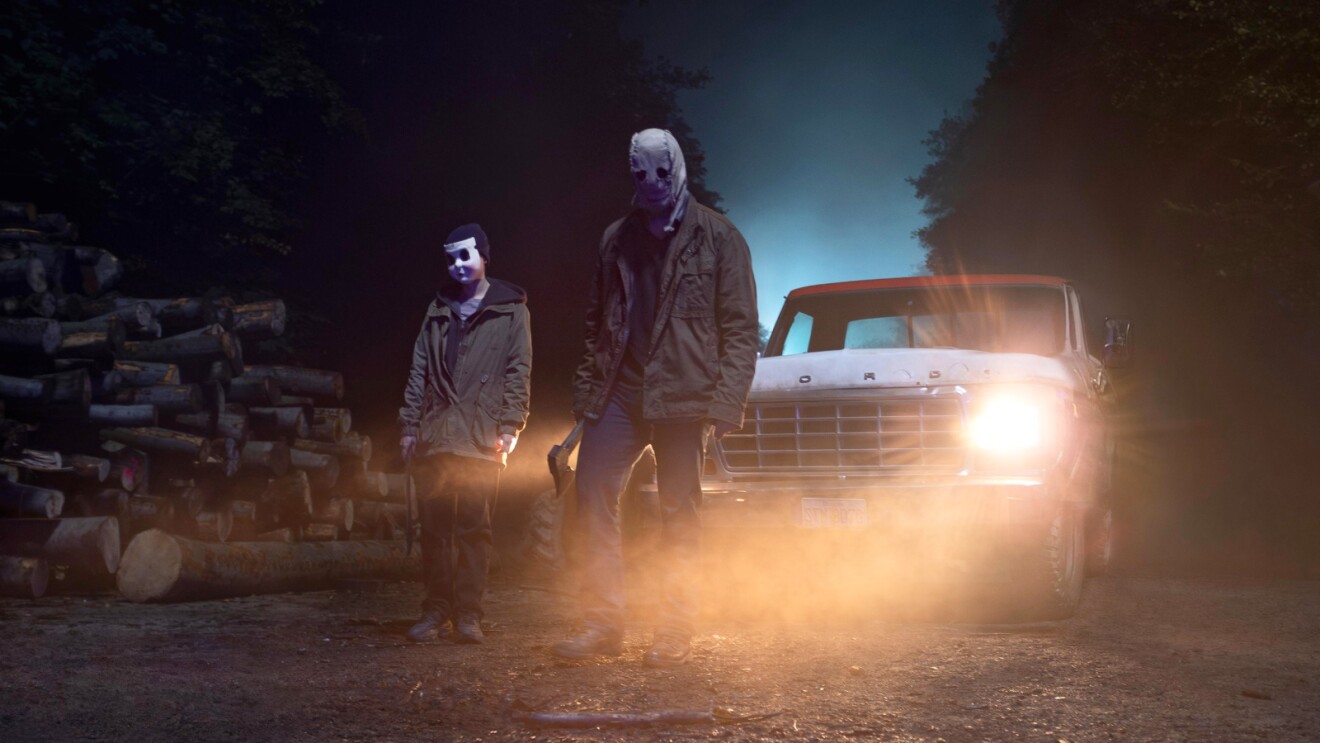 Two masked figures stand menacingly near a brightly lit truck at night