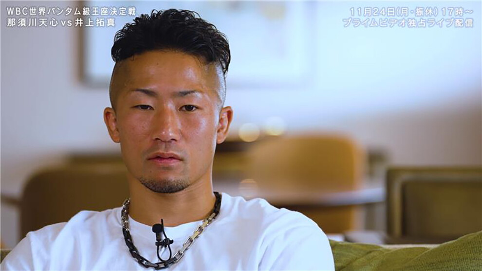 室内、ピントがぼやけた白い壁とテーブルや椅子などを背景に、男性のアップ。白いシャツでチェーン上のネックレスをつけ、襟元にマイクが取り付けられている。