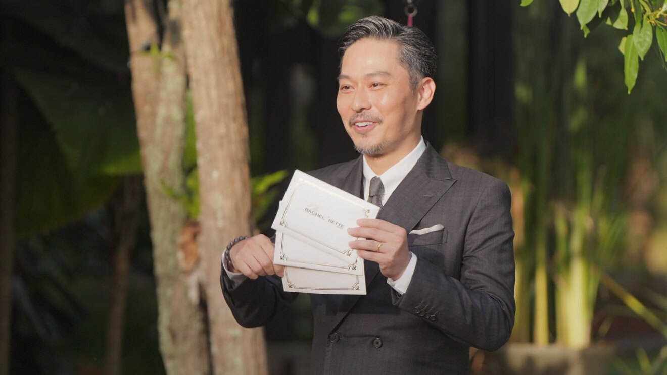 夜の屋外で、南洋の植物を背に立つスーツ姿の男性。体の前で、3つの封筒を持って見せている。