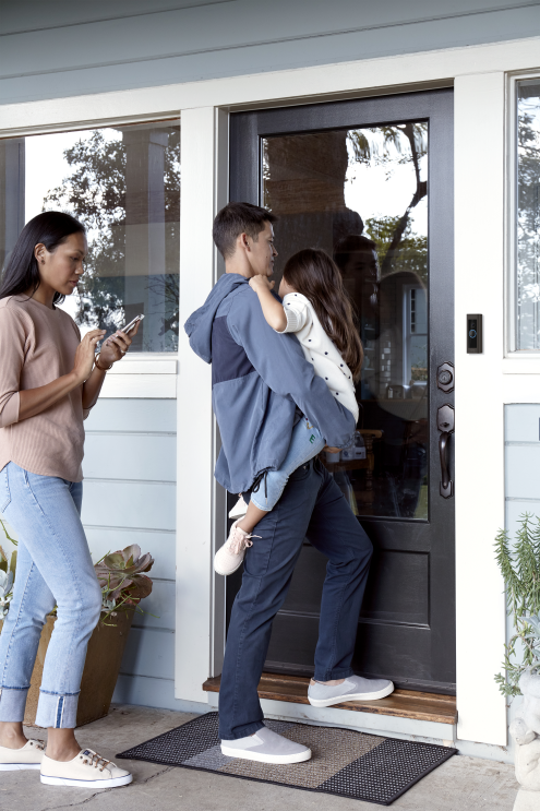 An image of a family walking into their house. A man is holding  a child while opening the door and a woman is close behind him using her phone.