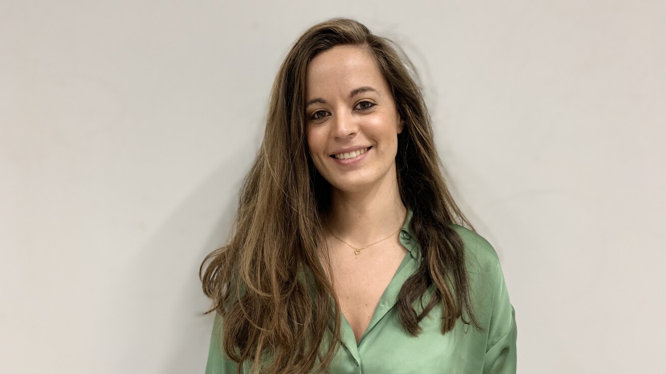 Eva. Pelo muy largo castaño claro con mechas rubias. Camisa escotada de seda verdes. Mira a casa sonriendo. De fondo, pared blanca.