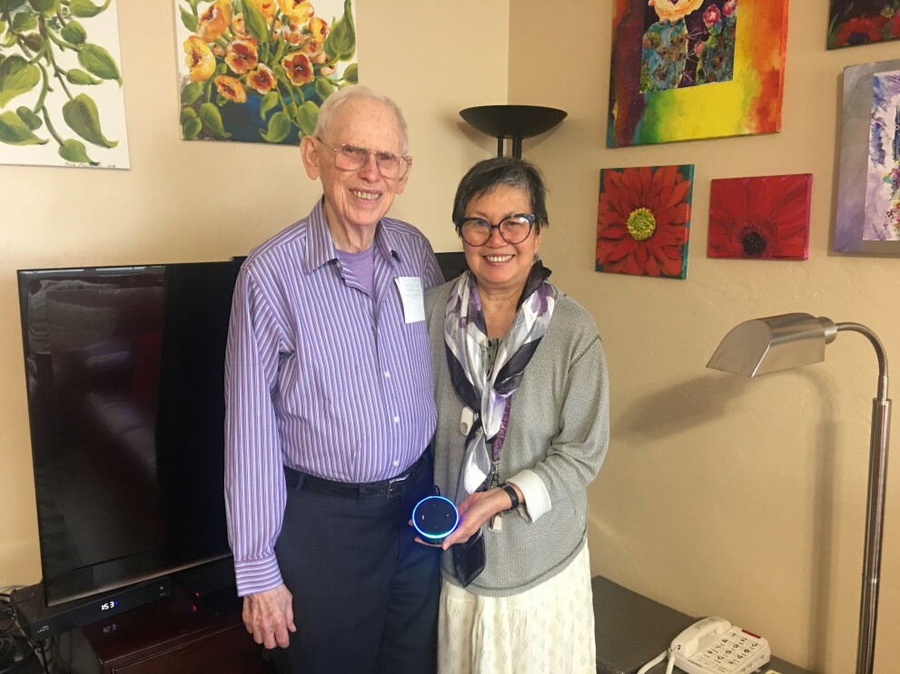 A man (Ron) and woman (Alicia) stand together in a living room, with colorful paintings on the wall behind him.