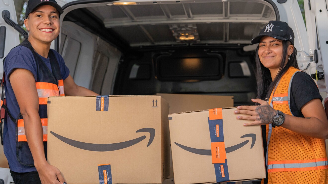 Dois entregadores, na frente de um furgão, segurando uma caixa cada. Eles estão de boné e usando coletes