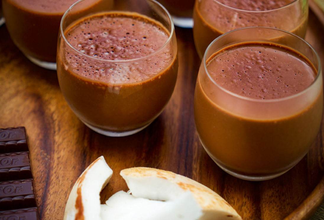 An image of chocolate pudding in small glasses sitting on a cutting board with chocolate and a slice of coconut. 
