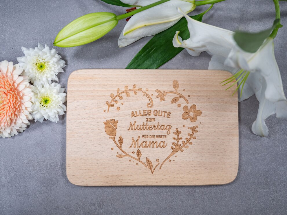 Auf einem grauen Tisch liegt ein Holzbrett mit der Aufschrift "Alles Gute zum Muttertag für die beste Mama"