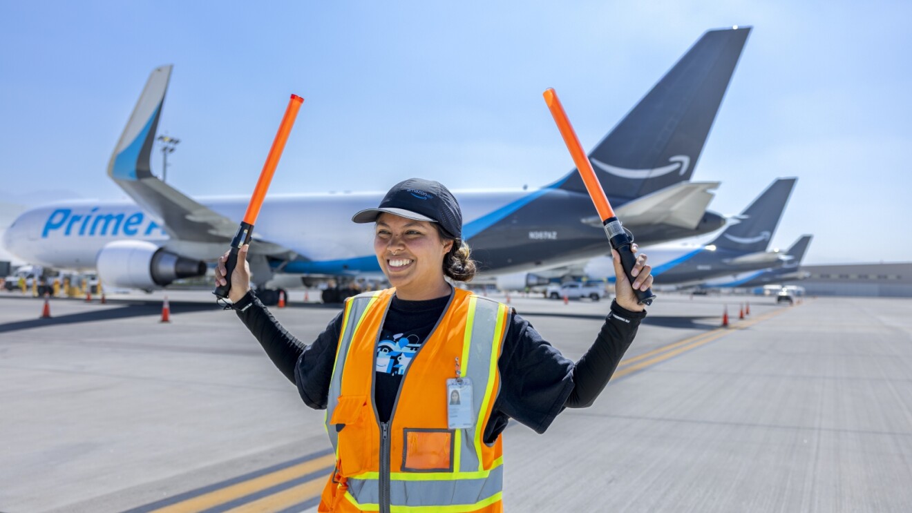 Smiling worker in safety vest directing aircraft on tarmac in front of Amazon Prime Air plane