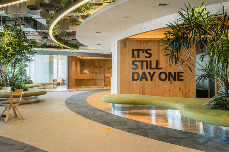 観葉植物が多数置かれた明るいオフィス入口。ウッディな壁にアルファベットの大きな文字で「IT'S STILL DAY ONE」と書かれている。