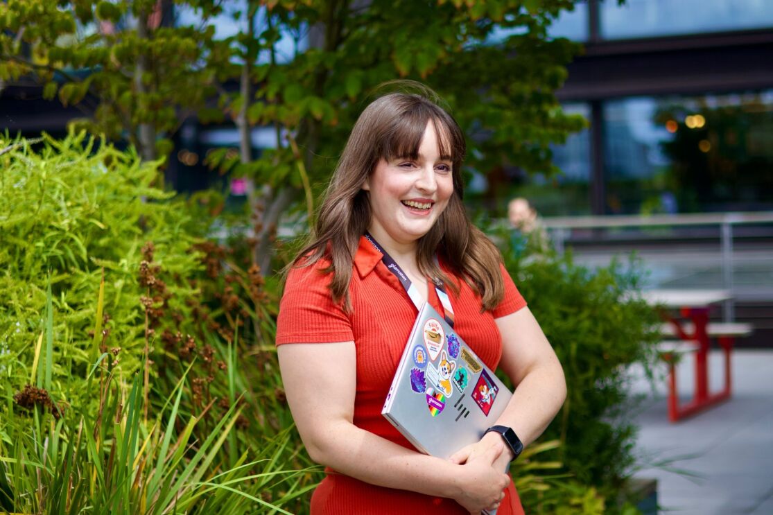 Smiling woman in red shirt holding laptop with stickers outdoors