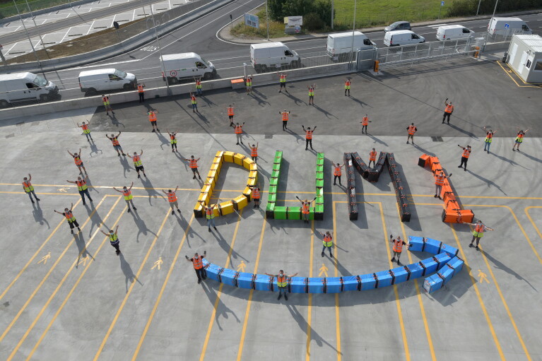 Foto aerea di un gruppo di dipendendi di un magazzino Amazon in Umbria posa con le braccia aperte e rivolte verso l'alto. A terra, alcuni sacchi colorati formano lo smile Amazon in azzurro e la scritta DUM1