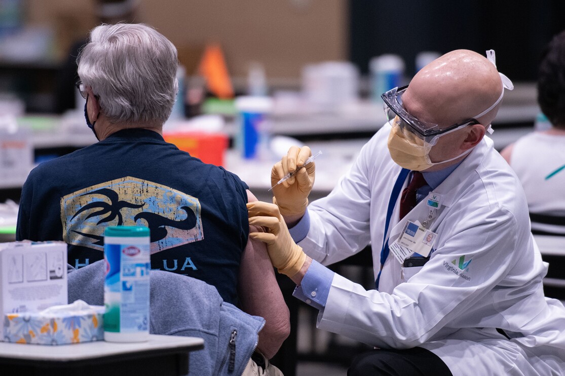 A man wearing a t-shirt receives a COVID-19 vaccination from a man wearing a Virginia Mason lab coat, mask, and gloves. 