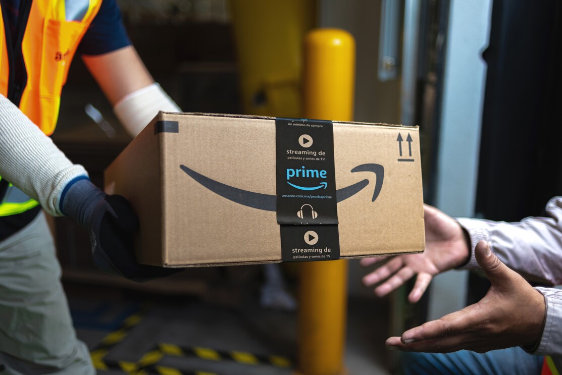 Primer plano de una caja de cartón de Amazon con las manos de un repartidor vestido con guantes negros y blanco u un chaleco de color naraja. Dos mano reciben la caja.