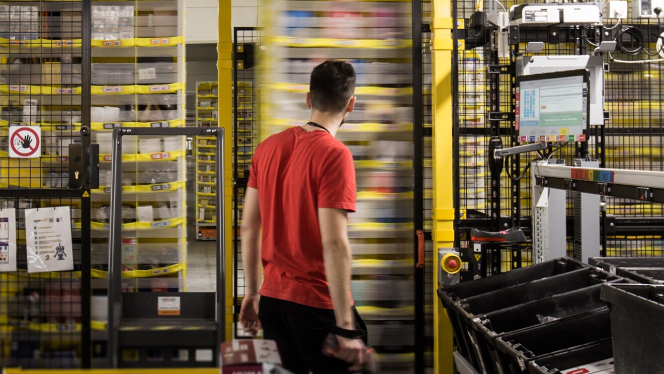 Un empleado de Amazon que se beneficia de nuestras estanterías móviles y unidades de transmisión. De fondo en el campo de las estanterías móviles, hay tres estanterías que se están moviendo y un empleado de espaldas que se lo mira. El empleado va con una camiseta de manga roja corta, un pantalón negro y en la mano derecha lleva el lector de código de barras.