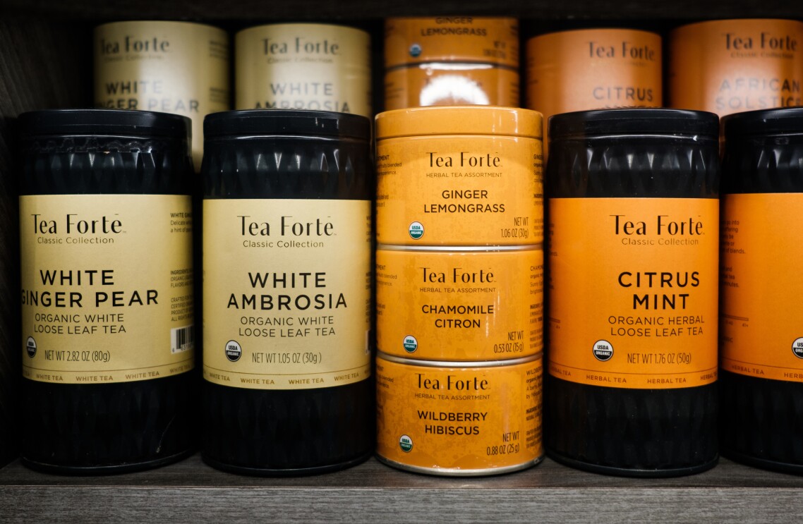 Canisters of Tea Forté tea sit on a shelf. Products shown include white ginger pear, white ambrosia, ginger lemongrass, chamomile citron, wildberry hibiscus, and citrus mint.