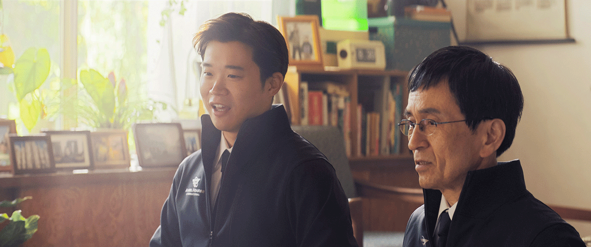 鉢植えや写真たてが多数飾られた部屋で、並んで座る2人の男性。揃いのジャケットとシャツを着用。左側の若い男性が話をしている。