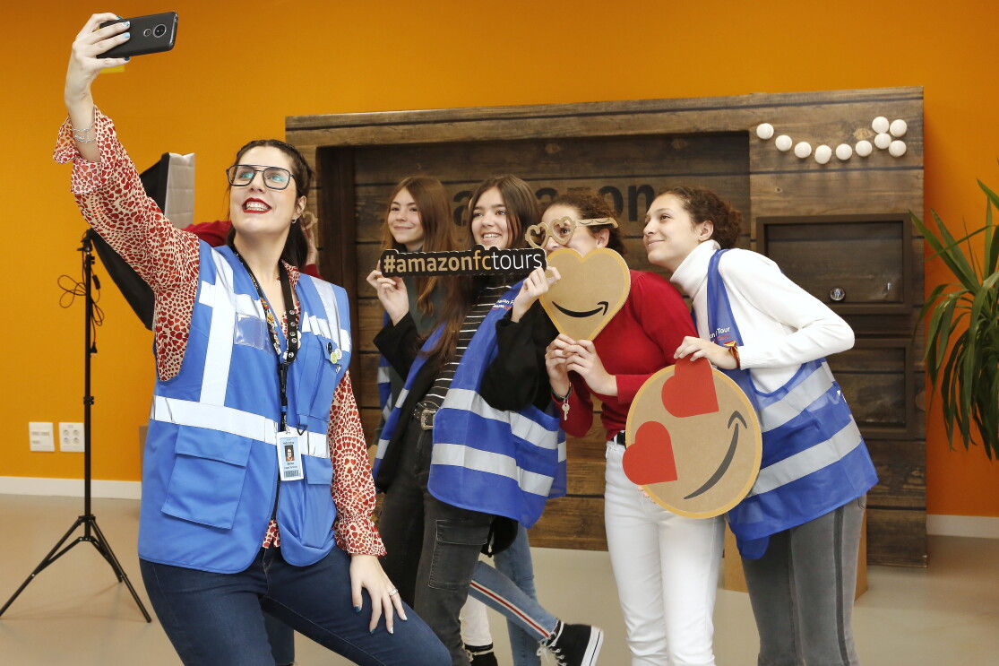 Delante de una decoración que simula una pared de madera con una luces de color y un foto, aparecen cinco mujeres haciéndose un selfie con un teléfono. Todas las protagonistas menos una lleva chaleco azul. De izquierda a derecha, primero la mujer que agarra el teléfono para el selfie. Lleva chaleco azul, una camisa estampada de animal print de color blanco y rojo y unos jeans. El pelo recogido y un badge colgando. La segunda mujer solo se le ve la cara y una melena larga de color castaño. La tercera mujer agarra un cartel que pone la etiqueta Amazon tours. Lleva el chaleco, unos jeans negros y un jersey de cuello alto con rayas. La cuarta mujer agarra una corazón con el logo de Amazon y unas gafas de cartón. La última mujer va con chaleco, un pantalón de color gris y un jersey blanco de cuello alto.