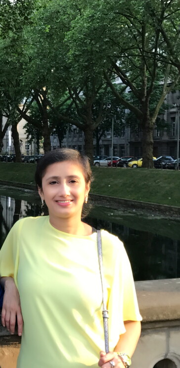 Ragini - wearing a neon yellow shirt and metallic purse leans against a ledge, smiling at the camera. Behind her are trees, and cars parked along a city street.