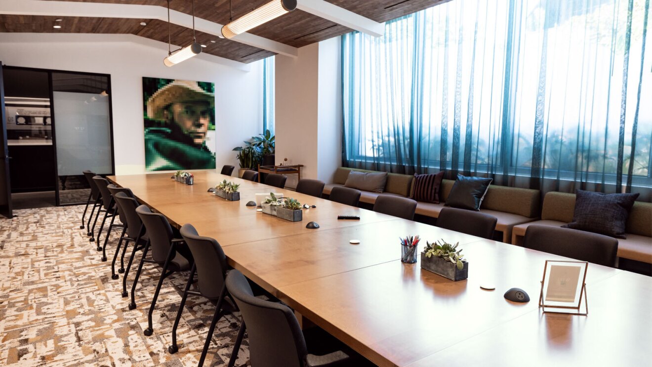 The boardroom inside the amazon music studio in los angeles. There is a large table with many chairs pulled up to it, beige carpet with a cool white design on it, exposed beams on the ceiling, and a digital art piece on the wall of a man in a cowboy hat.