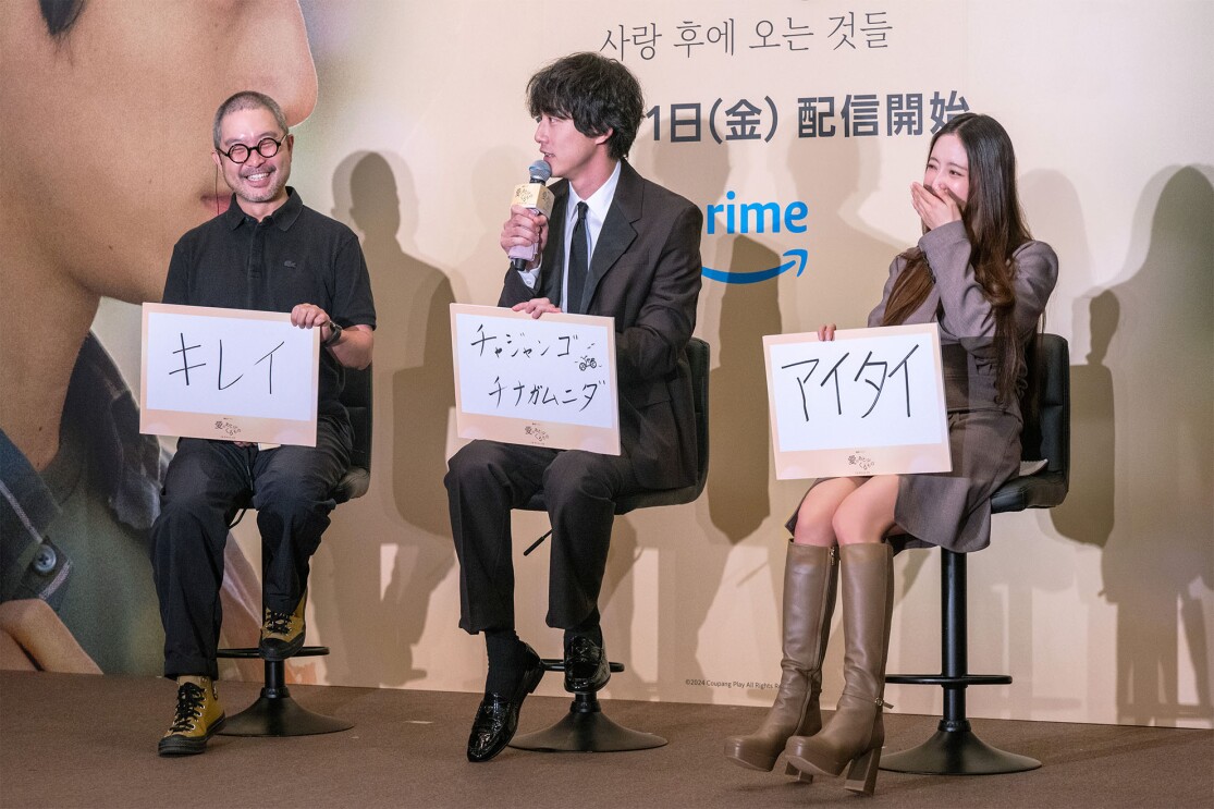 丸いメガネをかけ笑っている男性、マイクを持っている男性、口を手で隠している女性がそれぞれ文字が書かれたフラップを持ちハイチェアに座って並んでいる。