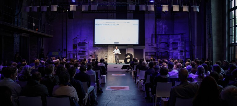 Ralf Kleber steht auf der Bühne der Amazon Academy 2017. Die Location ist voll mit Publikum und in der Mitte des Bilde sieht man den Mittelgang.