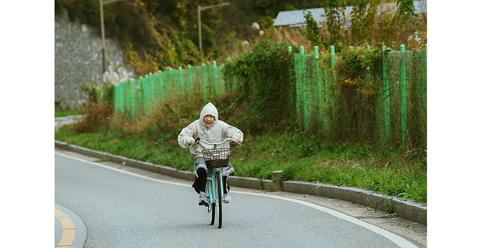 自転車をこいで舗装道路を走る人物。着用したパーカーのフードをかぶっている。道路の右側にはフェンスと背の高い植物やツタ。