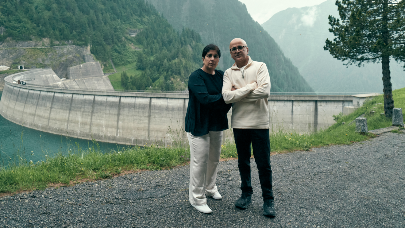 A middle-aged couple looking into camera infront of a dam and mountainous scenery