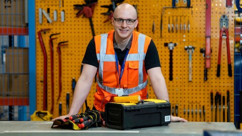 Luke Vials standing with his toolbox at his workbench wearing an orange high-viz vest.