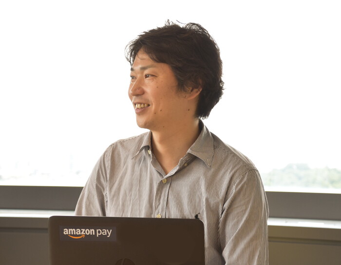 窓を背に話すグレーのシャツを着た男性。窓の外はほとんど白い空で、遠くに緑が見える。体の前に黒いノートパソコンを開いている。ノートパソコンの蓋に「amazon pay」の文字。