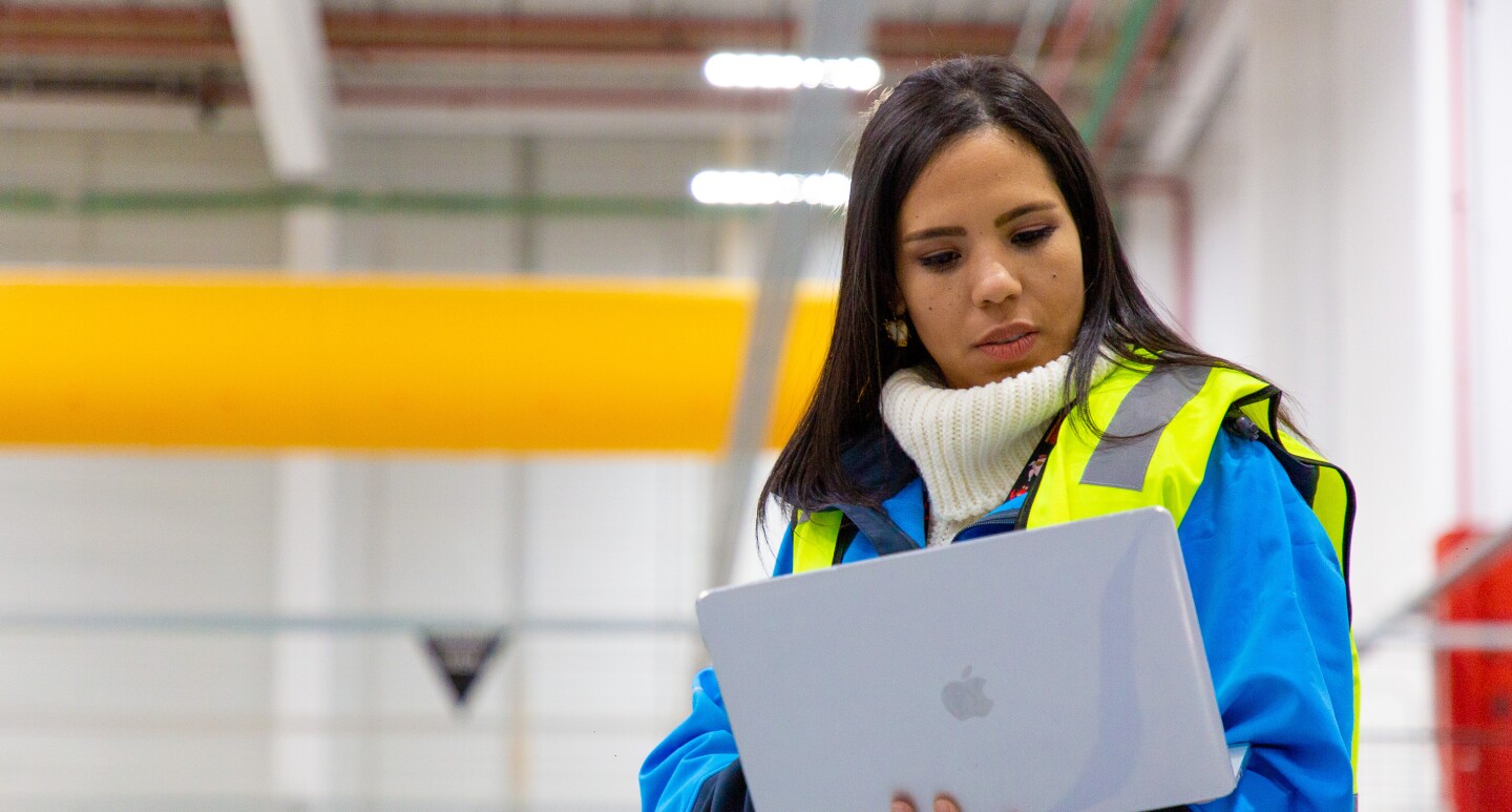 Ivette Duran, CEO de Cainaima Logistics, de pie con un ordenador entre las manos. De fondo está el centro logístico de color blanco y amarillo. Ella es morena y tiene el pelo largo y negro. Lleva un jersey de color blanco y de cuello alto, una chaqueta azul y un chaleco fluorescente.