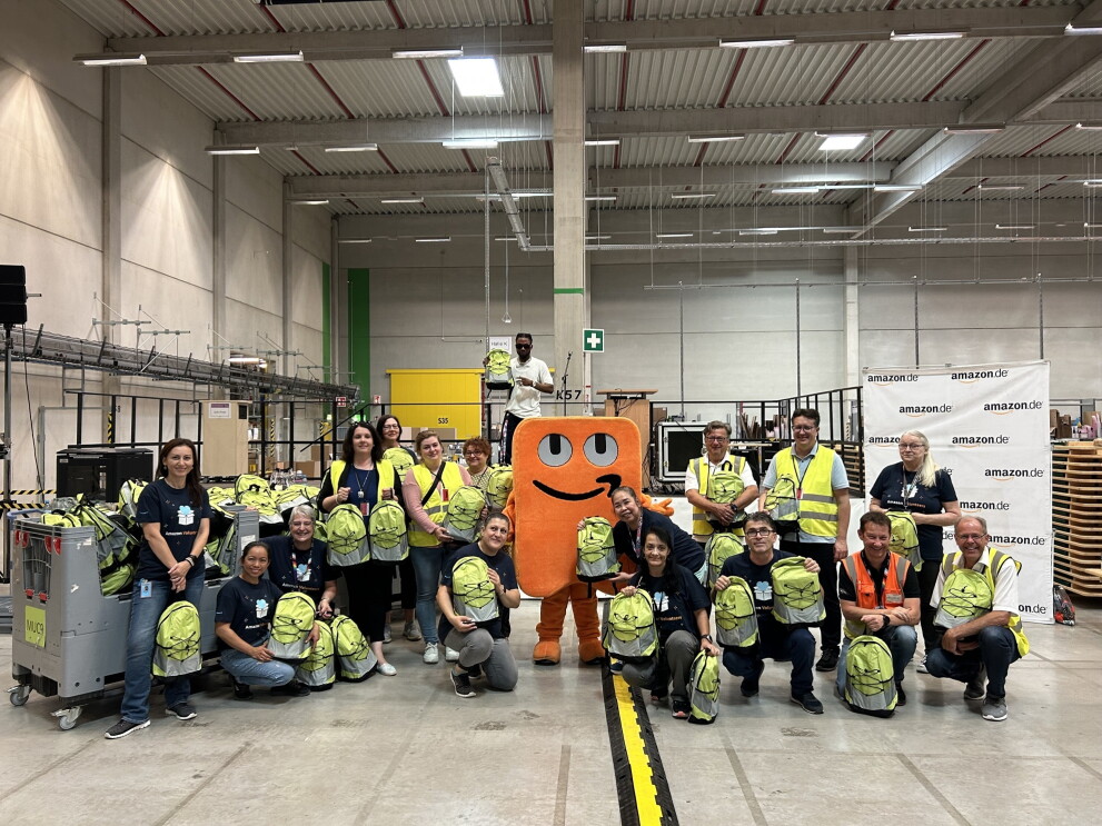 Amazon Mitarbeiterinnen und Mitarbeiter und Maskottchen Peccy halten im Logistikzentrum Graben bunte Back to School Rucksäcke