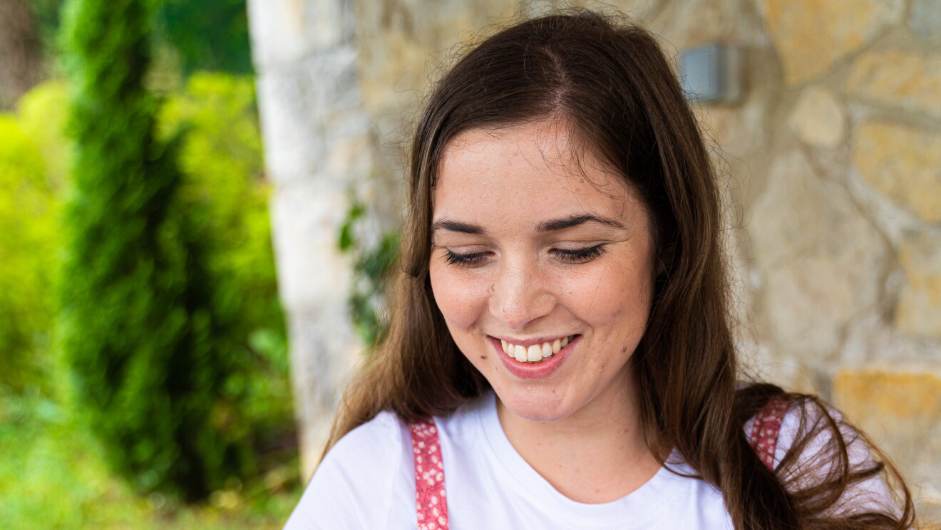 Alejandra Gómez Iribarnegaray tiene pelo largo y castaño. Lleva una camiseta de color blanco y encima un vestido de algodón con tirantes por encima de color blanco y rosa. 