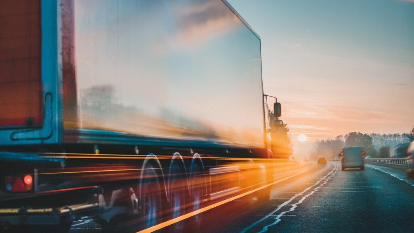 A semi truck drives down the highway with the sun setting in its path.