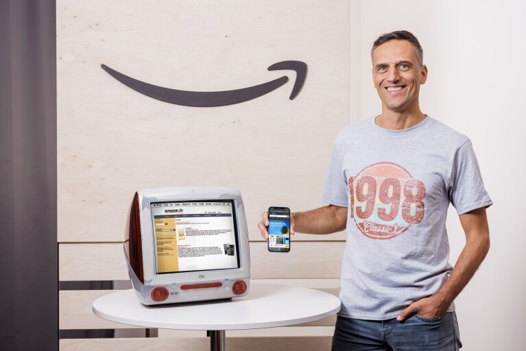 Amazon Deutschland gibt es seit 25 Jahren. Rocco Bräuniger hält ein Smartphone mit der Amazon App vor einen PC mit der Amazon Website.