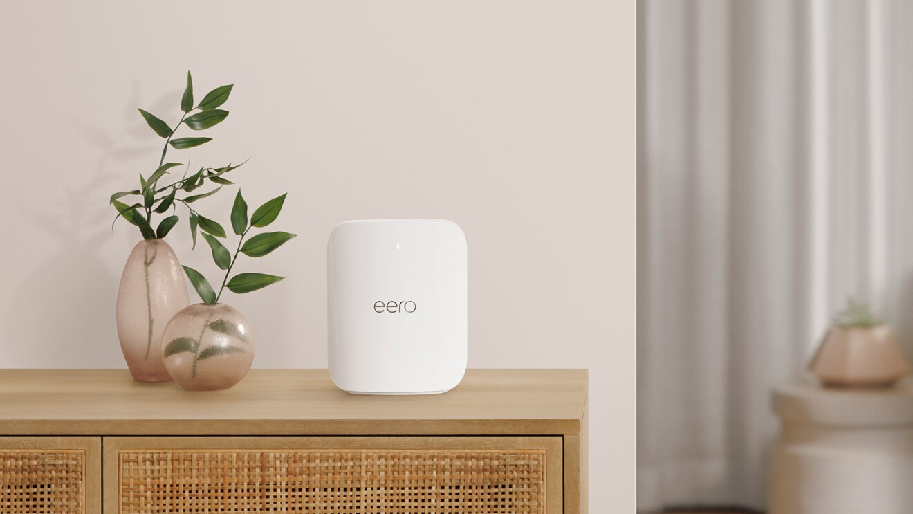 ベージュ色の壁の前に置かれた木製のラックの上に置かれた四角い白い機器(wifi)、その横に花瓶に入った観賞植物の葉