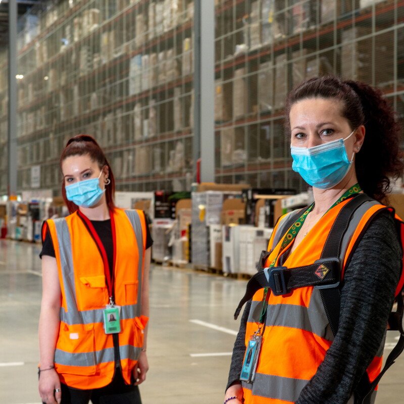 Two women, standing inside an Amazon fulfilment centre, wear orange safety vests and masks while looking at the camera.