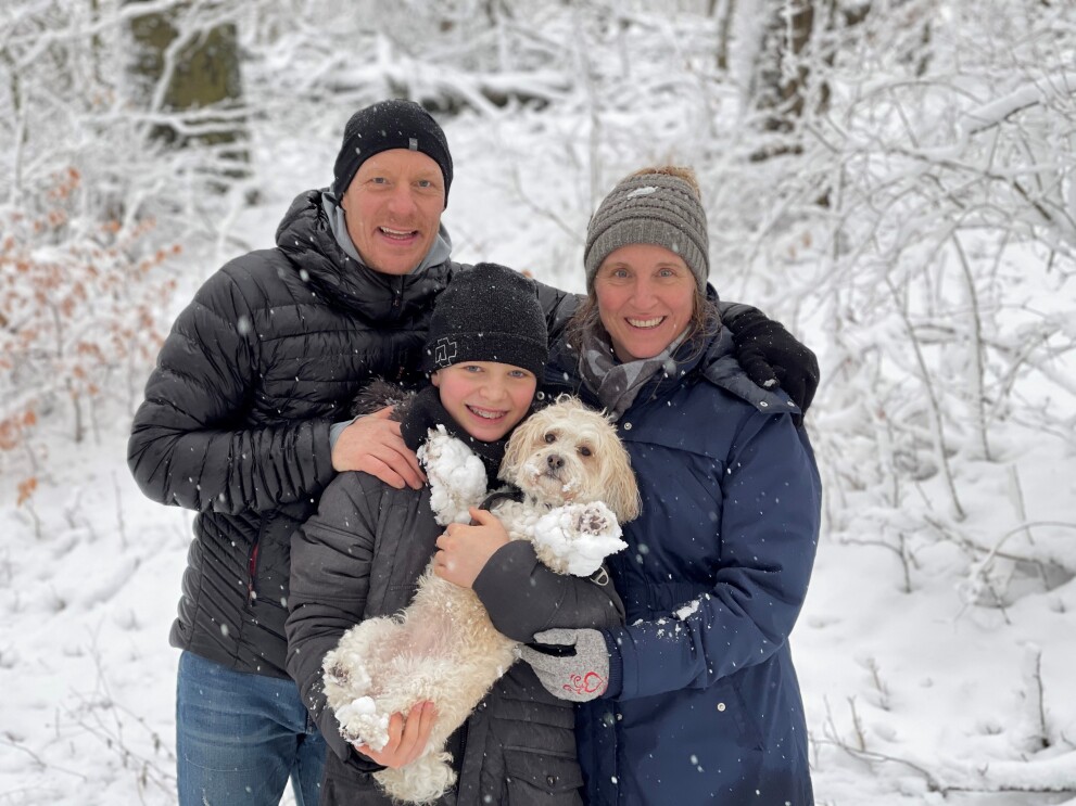Ein Mann, eine Frau und ein Kind mit Hund in den Armen stehen im Schnee und lachen in die Kamera.