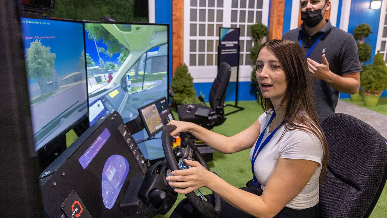 An image of a woman sitting in the driver's seat of an automated driving system. A man is standing behind her explaining the simulator.