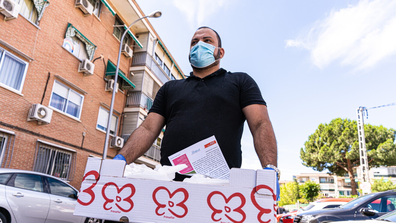 Yamil Pérez, colaborador de Amazon Flex. De fondo un edificio de 3 pisos de un barrio de clase trabajadora. Delante de los pisos coches aparcados y en primer plano el colaborador de Amazon Flex con guantes, mascarilla, una camiseta negra de manga corta, jeans y reloj. El pelo corto, barba negra. Lleva una caja de cartón blanca con flores rojas y se ven unas bolsas dentro. 
