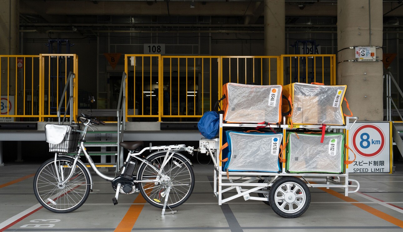 電動アシスト付き自転車の荷台が棚になっており、箱型の荷物を入れる袋が積まれている